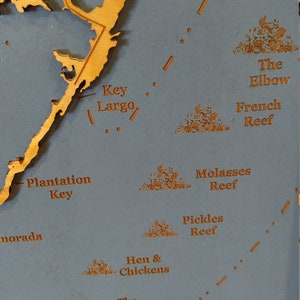 Puede incluir: Un primer plano de un mapa de madera de los Cayos de Florida, que muestra los nombres de varios arrecifes e islas, incluyendo Key Largo, Plantation Key y The Elbow. El mapa est&aacute; sobre un fondo azul.