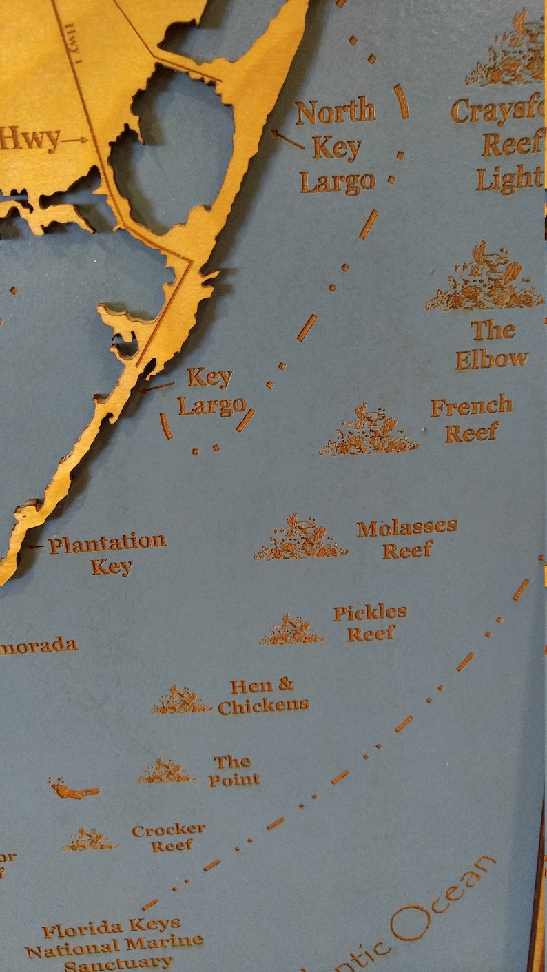 Puede incluir: Un primer plano de un mapa de madera de los Cayos de Florida, que muestra los nombres de varios arrecifes e islas, incluyendo Key Largo, Plantation Key y The Elbow. El mapa est&aacute; sobre un fondo azul.