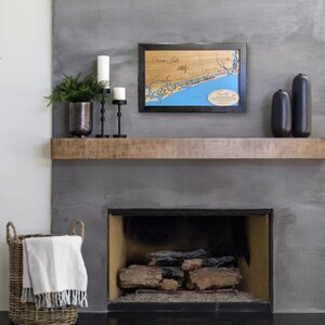Puede incluir: Un mapa de madera enmarcado de Ocean Isle, con agua azul y caracter&iacute;sticas terrestres marrones. El mapa se exhibe sobre una repisa de chimenea, junto con velas, jarrones decorativos y una cesta tejida con una manta.
