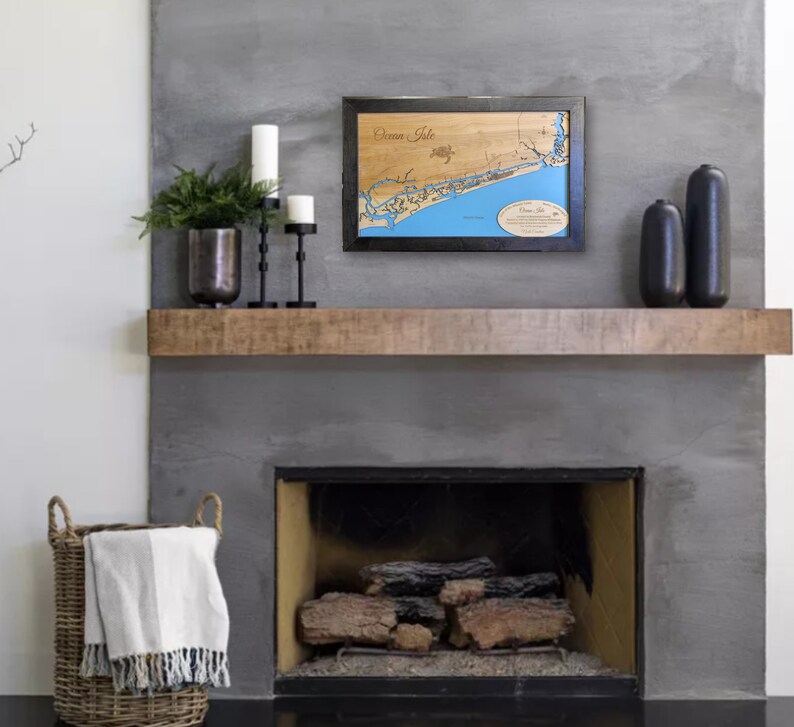 Puede incluir: Un mapa de madera enmarcado de Ocean Isle, con agua azul y caracter&iacute;sticas terrestres marrones. El mapa se exhibe sobre una repisa de chimenea, junto con velas, jarrones decorativos y una cesta tejida con una manta.