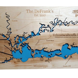 May include: A wooden map of the Tchoutacabouffa River in Mississippi, with the text "The DeFrank's Est. 2017" at the top. The river is outlined in blue, and the surrounding area is shown in brown. The map includes the names of various lakes and rivers, including Big Lake, Mullet Lake, and Biloxi River.