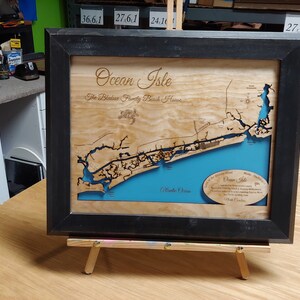 Puede incluir: Mapa enmarcado en madera de Ocean Isle, Carolina del Norte, con un marco negro. El mapa presenta una representaci&oacute;n detallada de la costa, con el oc&eacute;ano en azul y la tierra en tonos de madera natural. El texto incluye "Ocean Isle" y "The Bledsoe Family Beach House."