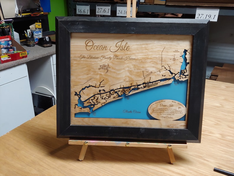 Puede incluir: Mapa enmarcado en madera de Ocean Isle, Carolina del Norte, con un marco negro. El mapa presenta una representaci&oacute;n detallada de la costa, con el oc&eacute;ano en azul y la tierra en tonos de madera natural. El texto incluye "Ocean Isle" y "The Bledsoe Family Beach House."