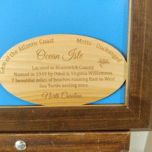 Puede incluir: Placa de madera con el texto "Gem of the Atlantic Coast Ocean Isle" y otros detalles sobre la ubicaci&oacute;n, incluyendo "North Carolina". La placa est&aacute; colocada sobre un fondo azul dentro de un marco de madera oscura.
