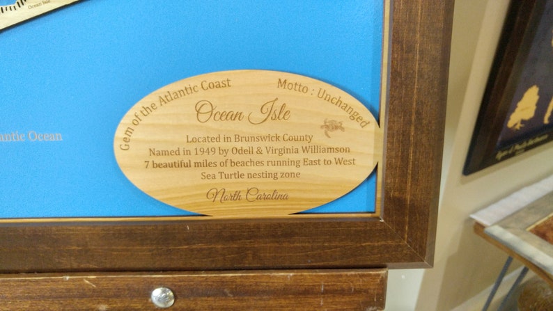 Puede incluir: Placa de madera con el texto "Gem of the Atlantic Coast Ocean Isle" y otros detalles sobre la ubicaci&oacute;n, incluyendo "North Carolina". La placa est&aacute; colocada sobre un fondo azul dentro de un marco de madera oscura.