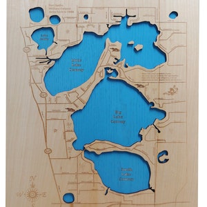 May include: A wooden map of the Conway Chain of Lakes in Florida. The map shows the four interconnected lakes: East, West, Middle, and South. The map is laser-cut and painted blue to represent the water. The map is mounted on a wooden plaque.