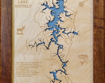May include: A wooden map of Lake Nottely in Georgia, with a blue lake and brown land. The map is framed in a dark brown wood frame. The text "LAKE NOTTELY" is at the top of the map. The text "GEORGIA" is at the bottom of the map. The text "Lake Nottely is one of many reservoirs of the Tennessee Valley Authority. It is located in Union County, Georgia. Formed in 1942 by the damming of the Nottely River. Lake Nottely extends 20 miles upstream to the town of Blairsville. 4,180 surface acres at normal summer pool elevations." is on the right side of the map.