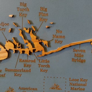 Puede incluir: Un mapa de madera de los Cayos de Florida con los nombres de varias islas y puntos de referencia etiquetados. El mapa est&aacute; sobre un fondo azul y presenta un acabado de madera marr&oacute;n claro.