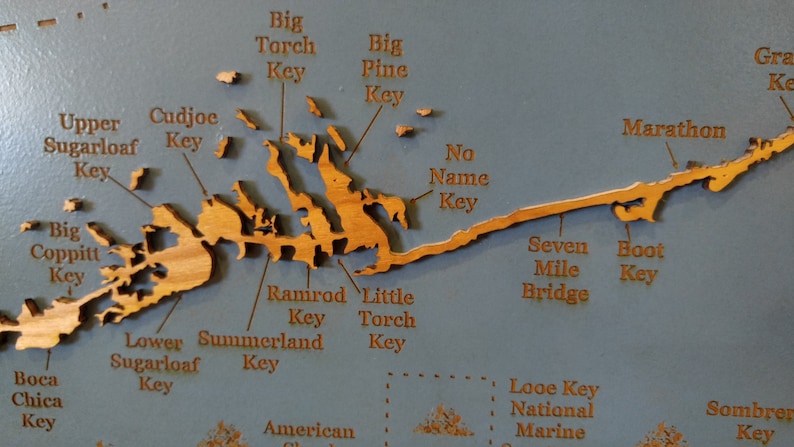 Puede incluir: Un mapa de madera de los Cayos de Florida con los nombres de varias islas y puntos de referencia etiquetados. El mapa est&aacute; sobre un fondo azul y presenta un acabado de madera marr&oacute;n claro.