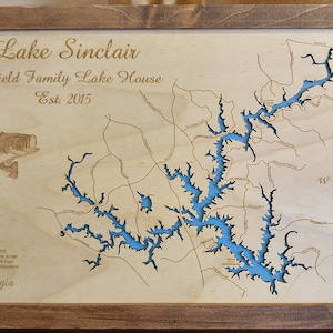 May include: Wooden framed map of Lake Sinclair with blue water detail. The map includes the text "Lake Sinclair", "Barfield Family Lake House", and "Est. 2015". A compass rose is in the upper right corner. A fish graphic is in the upper left corner.