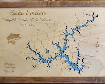 May include: Wooden framed map of Lake Sinclair with blue water detail. The map includes the text "Lake Sinclair", "Barfield Family Lake House", and "Est. 2015". A compass rose is in the upper right corner. A fish graphic is in the upper left corner.
