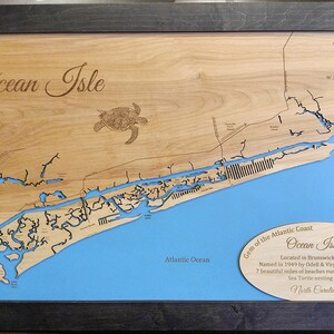 Puede incluir: Mapa de madera de Ocean Isle, Carolina del Norte, con un fondo azul que representa el Oc&eacute;ano Atl&aacute;ntico. El mapa est&aacute; cortado con l&aacute;ser con detalles intrincados, incluyendo una tortuga y el texto "Ocean Isle". El mapa est&aacute; enmarcado en un marco de madera gris oscuro.