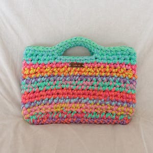 May include: A rectangular crochet handbag featuring horizontal stripes in vibrant shades of mint green, bright pink, yellow, and light purple. The bag has integrated handles and a small rectangular metal tag centered on the front with the text Lovelle Lane.