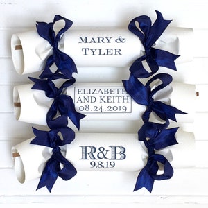 May include: Four white paper party poppers with blue ribbon bows. The poppers are decorated with the names of couples and their wedding dates. The text on the poppers reads: "Mary & Tyler", "Elizabeth and Keith 08.24.2019", "R&B 9.8.19".