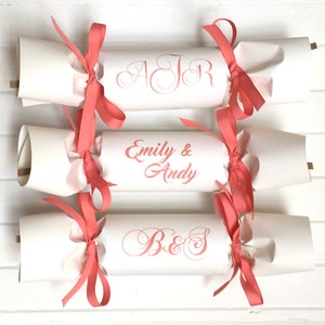 May include: Three white paper Christmas crackers with coral pink ribbon bows. The crackers are decorated with personalized text in coral pink lettering. The text on the crackers reads "AJR", "Emily & Andy", and "B&S".