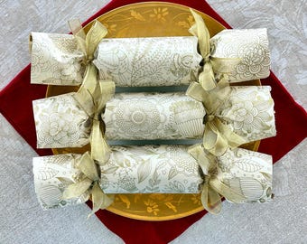 May include: Three cylindrical party crackers wrapped in cream-colored paper with a gold floral and bird line-art pattern. Each cracker is tied at both ends with a sheer gold ribbon, creating a gathered effect on the paper at the closure points.