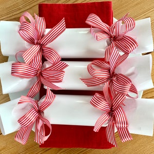 May include: A festive arrangement of white paper crackers tied with red and white striped ribbons. The crackers are arranged on white plates, with a red napkin visible in the background. The scene is set on a light wood surface.