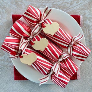 May include: Three cylindrical party crackers wrapped in red and white striped paper, tied at both ends with striped fabric ribbons. Each cracker features a plain, light-colored wooden heart-shaped tag attached to the center ribbon.