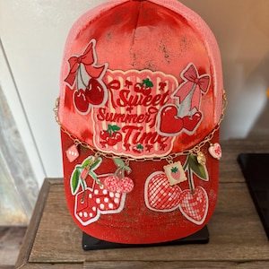 May include: A red and pink trucker hat featuring a central patch with the text Sweet Summer Time, surrounded by embroidered cherry, bow, and dice motifs. A gold-toned chain drapes across the front, adorned with small charms including hearts and fruit.