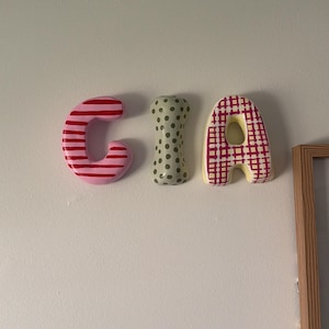 May include: Three-dimensional wall letters spelling the name GIA. The letter G features horizontal pink and red stripes. The letter I is light green with dark green polka dots. The letter A has a yellow base with a red grid pattern overlay.