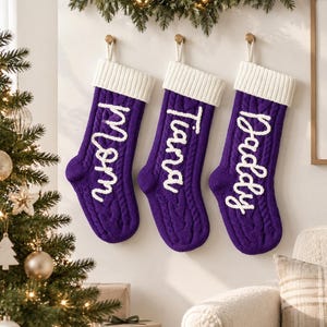 Puede incluir: Tres calcetines navideños de punto trenzado en color morado con puños acanalados blancos. Cada calcetín presenta un nombre bordado en letra cursiva blanca en la parte frontal, Mom, Tiara y Daddy, colgados de ganchos en la pared.