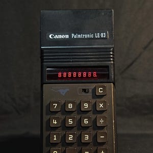 Puede incluir: Una calculadora de bolsillo negra Canon Palmtronic LE-83 con una pantalla LED roja que muestra ocho dígitos. El dispositivo tiene un cuerpo rectangular con un teclado numérico, teclas de función y un interruptor sobre los botones numéricos.