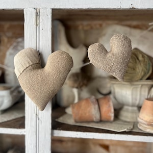May include: Two heart-shaped burlap ornaments in a tan colour. One has a contrasting fabric detail. They hang in front of a white cabinet, with other decorative items visible inside. The cabinet has a distressed finish.