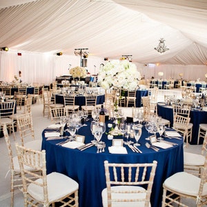 Puede incluir: Un lugar de recepción de boda decorado con cortinas de tela blanca y centros de mesa florales blancos. Las mesas están cubiertas con manteles azul marino y tienen sillas doradas.