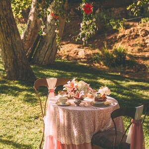 May include: A pink tablecloth with lace overlay covers a round table set for two in a garden setting. The table is decorated with a bouquet of white and pink flowers, and there are two chairs with pink sashes.
