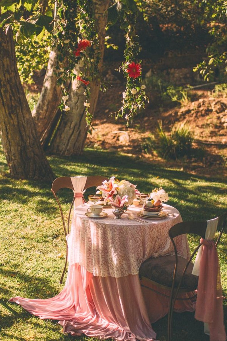 May include: A pink tablecloth with lace overlay covers a round table set for two in a garden setting. The table is decorated with a bouquet of white and pink flowers, and there are two chairs with pink sashes.