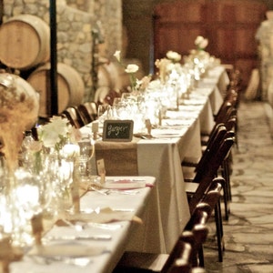 May include: A long, white tablecloth with a burlap runner is set with candles, glasses, and place cards. The table is surrounded by wooden chairs and is lit by string lights hanging from the ceiling.