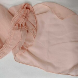 May include: A peach-colored sheer fabric scarf with a ruffled edge. The scarf is folded in half and the edges are sewn together.