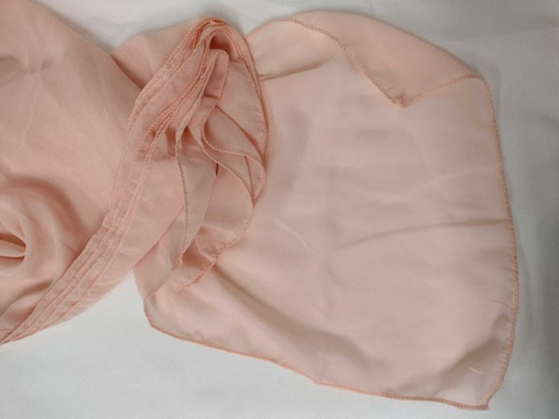 May include: A peach-colored sheer fabric scarf with a ruffled edge. The scarf is folded in half and the edges are sewn together.