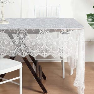 May include: White lace tablecloth with a scalloped edge. The tablecloth is draped over a wooden table with two white chairs.