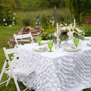 Puede incluir: Una mesa blanca con un mantel con volantes preparada para una boda o un evento especial. La mesa está decorada con vasos verdes, flores blancas y velas. Dos sillas blancas se colocan a cada lado de la mesa.