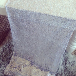 May include: A silver and gold sequined table runner with a gradient design. The runner is made of small, shiny sequins that create a shimmering effect.