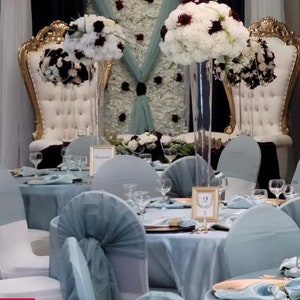 K&ouml;nnte beinhalten: Ein Hochzeitsempfang-Tisch mit wei&szlig;en und burgunderfarbenen Blumenarrangements in hohen Glasvasen. Die Tische sind mit hellblauen Tischdecken bedeckt und die St&uuml;hle mit hellblauem Stoff bezogen.
