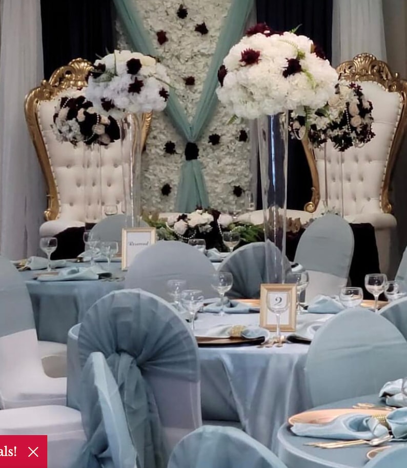 K&ouml;nnte beinhalten: Ein Hochzeitsempfang-Tisch mit wei&szlig;en und burgunderfarbenen Blumenarrangements in hohen Glasvasen. Die Tische sind mit hellblauen Tischdecken bedeckt und die St&uuml;hle mit hellblauem Stoff bezogen.