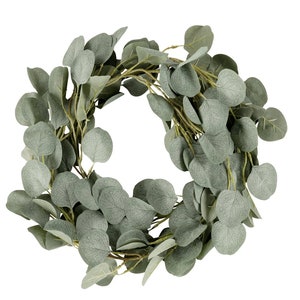 Puede incluir: Una corona de eucalipto verde hecha de hojas artificiales. La corona es redonda y tiene un diámetro de unos 30 cm.