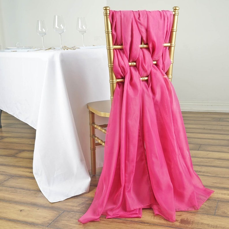 May include: A bright pink chair sash tied in a decorative knot pattern on the back of a gold chair. The sash is made of a sheer fabric and drapes down the back of the chair.