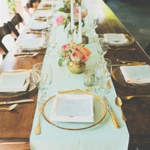 May include: A light blue table runner with gold accents is set for a wedding reception. The table is decorated with white plates, gold cutlery, and clear glasses. A vase of pink and white flowers sits in the center of the table.