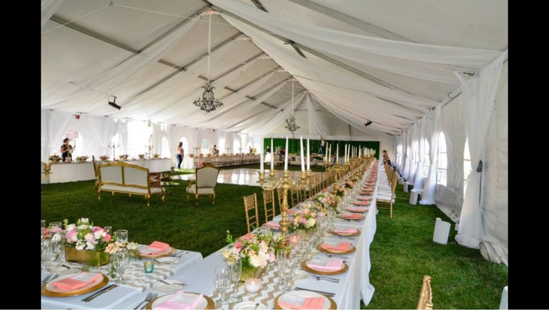 Puede incluir: Una larga carpa blanca con tela blanca que cae del techo. La carpa est&aacute; decorada con candelabros dorados y flores. Hay mesas dispuestas con manteles blancos y servilletas rosas. Las mesas est&aacute;n rodeadas de sillas doradas.