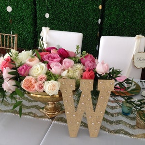 Puede incluir: Una mesa de boda con un centro de mesa de rosas rosas y blancas en un jarr&oacute;n dorado. La mesa est&aacute; decorada con un camino de mesa de chevron dorado y blanco y una letra W de purpurina dorada. Un cartel que dice "Bride" est&aacute; en una silla blanca.