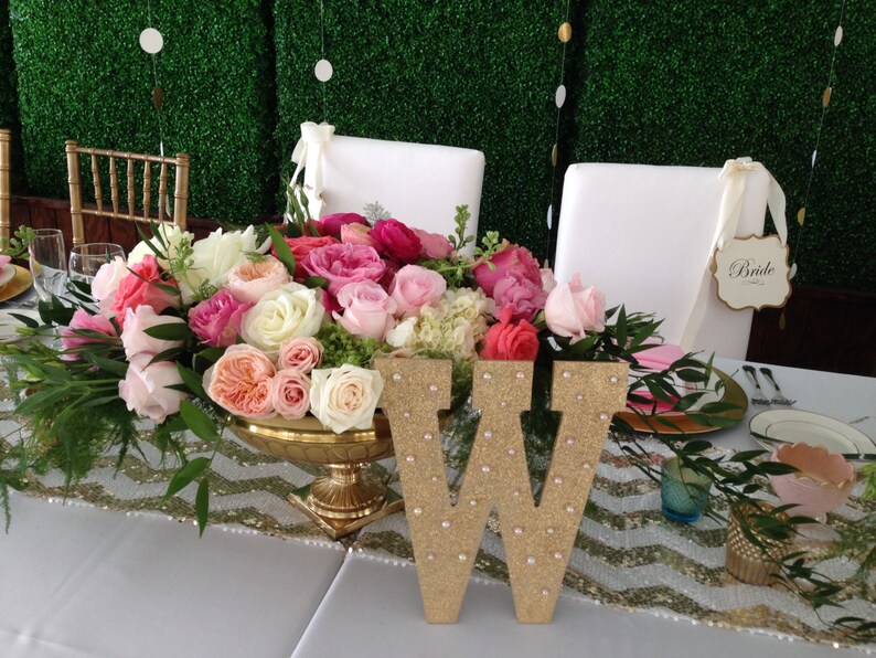 Puede incluir: Una mesa de boda con un centro de mesa de rosas rosas y blancas en un jarr&oacute;n dorado. La mesa est&aacute; decorada con un camino de mesa de chevron dorado y blanco y una letra W de purpurina dorada. Un cartel que dice "Bride" est&aacute; en una silla blanca.