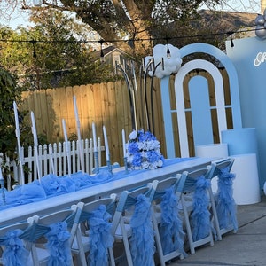 K&ouml;nnte beinhalten: Ein wei&szlig;er Tisch mit blauen, gekr&auml;uselten Stuhlb&auml;ndern ist im Freien f&uuml;r eine Party aufgebaut. Der Tisch ist mit wei&szlig;en Kerzen und einem Mittelst&uuml;ck aus wei&szlig;en und blauen Blumen dekoriert. Im Hintergrund ist eine blaue und wei&szlig;e Torbogen-Kulisse zu sehen.
