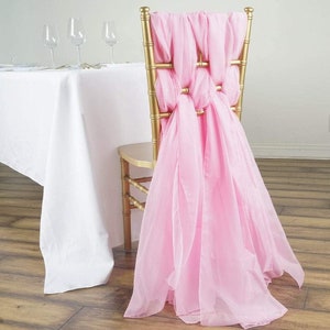 May include: Pink chiffon chair sash tied in a decorative knot on a gold chair. The sash is draped over the back of the chair and hangs down to the floor.