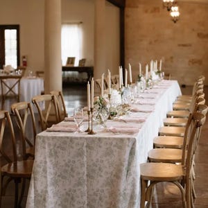 Pode incluir: Uma longa toalha de mesa branca com padrão floral está preparada para uma recepção de casamento. A mesa é decorada com velas brancas e flores. As cadeiras são de madeira com encosto cruzado.
