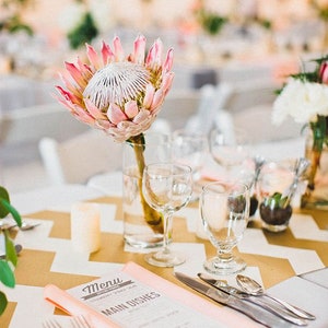 Puede incluir: Una mesa de boda con una flor de protea rosa y blanca en un jarrón, una tarjeta de menú con las palabras "Menú" y "Platos principales", y un mantel con estampado de chevron dorado y blanco.