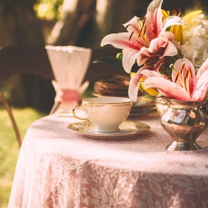 May include: A pink lace tablecloth with a white teacup and saucer, a silver creamer, and a bouquet of pink lilies.