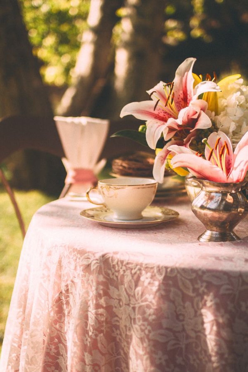 May include: A pink lace tablecloth with a white teacup and saucer, a silver creamer, and a bouquet of pink lilies.